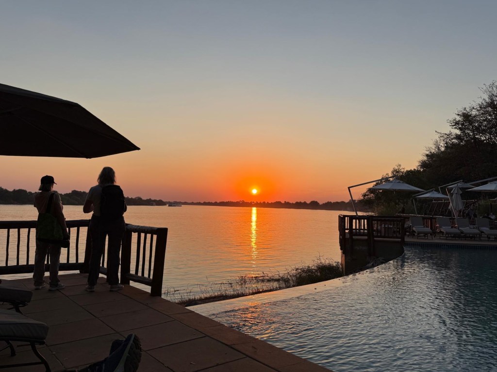 Two people standing by a poolside overlooking a sunset by the water, with silhouettes visible against the colorful sky.